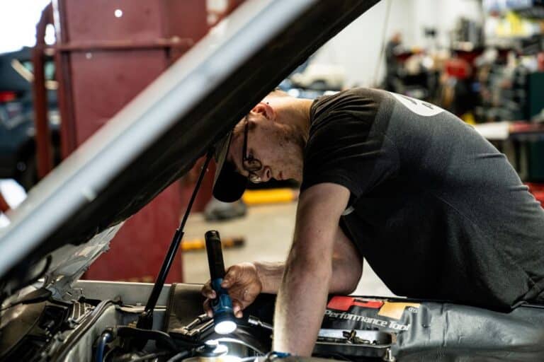 Auto repair technician using flashlight to look under car hood.